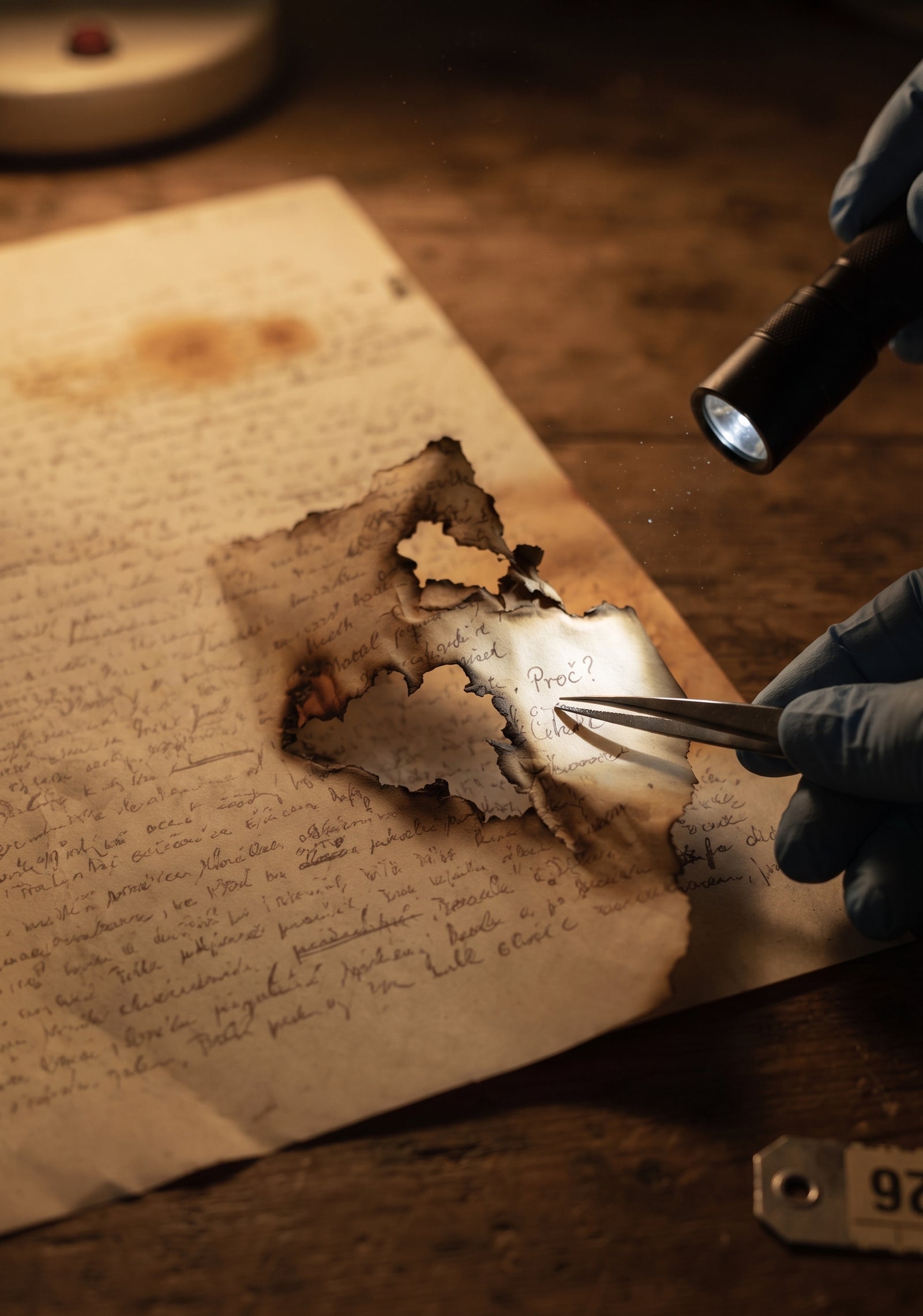 A gloved hand examines a burned letter with tweezers and a flashlight, the word 'Proc?' visible through a scorched hole in the page
